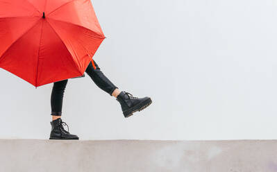 Anonymous young female with red umbrella walking and balancing on border against gray wall on rainy day on street - ADSF29411