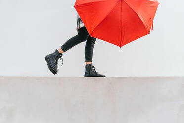 Anonymous young female with red umbrella walking and balancing on border against gray wall on rainy day on street - ADSF29412