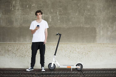 Teenage boy listening music while standing by electric push scooter at basement - MAEF13052