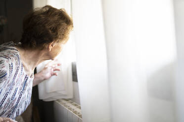 Senior woman looking through window at home - JCCMF03768