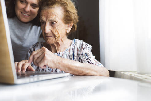 Lächelnde junge Frau, die ihrer Großmutter bei der Benutzung eines Laptops zu Hause hilft - JCCMF03775