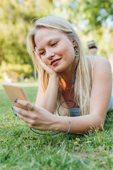 Smiling charming female lying on grass in park and listening to music in headphones in summer - ADSF29433