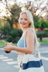 Side view of cheerful female standing with cold lemonade in plastic cup in street in summer looking at camera - ADSF29436