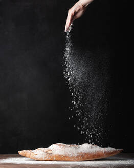 Crop anonymous baker sprinkling white flour over freshly baked artisan baguette bread against black background - ADSF29438