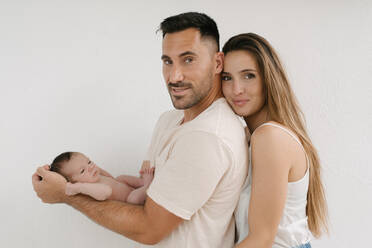 Side view of delighted couple with naked infant looking at camera on white background - ADSF29446