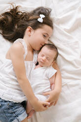 Draufsicht auf ein fröhliches kleines Mädchen, das ein bezauberndes Kleinkind umarmt, während es auf einem weichen Bett zu Hause liegt - ADSF29453