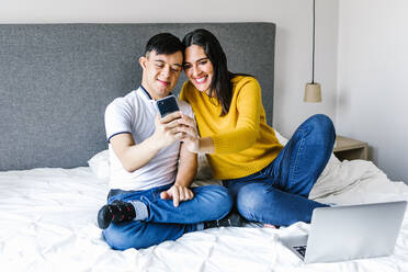Erfreute ethnische Mutter und Teenager mit Down-Syndrom sitzen auf dem Bett und machen ein Selbstporträt mit dem Smartphone - ADSF29474