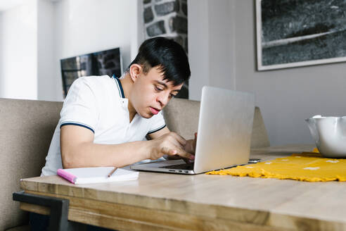 Fokus Lateinischer Teenager mit Down-Syndrom, der am Tisch sitzend auf seinem Netbook surft und von zu Hause aus online lernt - ADSF29475