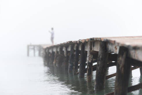 Gesichtslose Person steht auf einem Holzsteg in der Nähe des Meeres am Playa de Muro und genießt den nebligen Morgen - ADSF29477