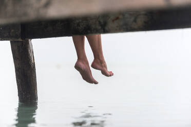 Crop legs of female sitting on wooden quay near sea on misty morning on Playa de Muro - ADSF29478