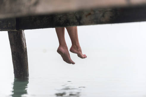 Crop Beine der Frau sitzen auf hölzernen Kai in der Nähe von Meer auf nebligen Morgen auf Playa de Muro - ADSF29478