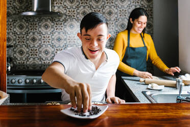 Begeisterter ethnischer Teenager mit Down-Syndrom verziert Kekse mit Schokoladenstückchen, während er mit seiner lächelnden Mutter zu Hause Gebäck zubereitet - ADSF29482