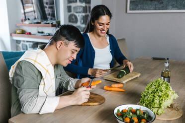 Fröhliche ethnische Mutter und Sohn im Teenageralter mit Down-Syndrom sitzen am Tisch und schneiden Gemüse, während sie Salat für das Mittagessen zu Hause vorbereiten - ADSF29483