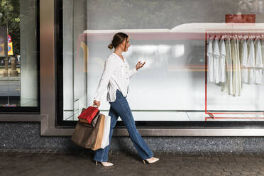 Young woman using smart phone while walking with shopping bags by store - JRVF01733