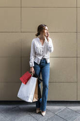 Smiling woman with shopping bags talking on smart phone in front of wall - JRVF01738