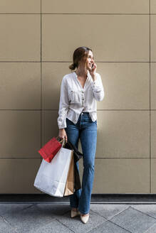 Lächelnde Frau mit Einkaufstüten, die vor einer Wand mit ihrem Smartphone telefoniert - JRVF01738