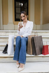 Woman using mobile phone while sitting with shopping bags on staircase - JRVF01745