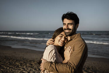 Happy young couple embracing while standing at beach - RCPF01213