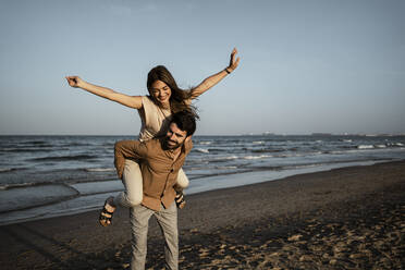 Playful woman with arms outstretched enjoying piggyback on boyfriend during sunset - RCPF01220