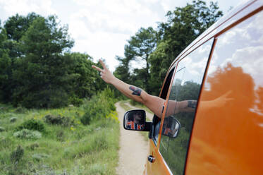 Crop anonymous traveler driving van on road in forest and showing peace sign during summer trip - ADSF29508