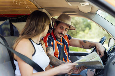 Side view of couple of travelers sitting in van and orientating with paper map during road trip in summer - ADSF29512