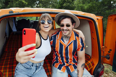 Vergnügtes Paar von Reisenden, die in einem Van sitzen und sich mit ihrem Smartphone fotografieren, während sie sich umarmen und das Sommerabenteuer genießen - ADSF29520