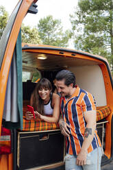 Vergnügtes Paar von Reisenden, die in einem Van sitzen und sich mit ihrem Smartphone fotografieren, während sie sich umarmen und das Sommerabenteuer genießen - ADSF29524