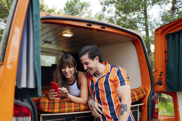 Vergnügtes Paar von Reisenden, die in einem Van sitzen und sich mit ihrem Smartphone fotografieren, während sie sich umarmen und das Sommerabenteuer genießen - ADSF29525
