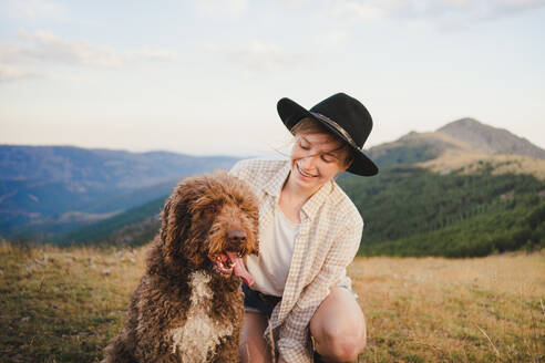 Fröhliche Besitzerin mit gehorsamem Labradoodle-Hund sitzt in den Bergen und schaut weg - ADSF29533