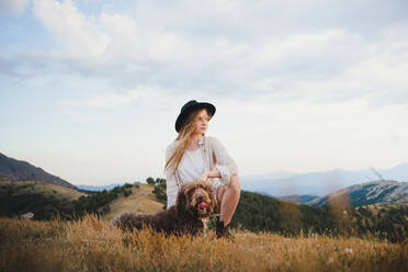 Female owner with obedient Labradoodle dog sitting in mountains looking away - ADSF29534