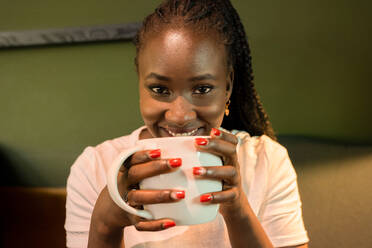 Positive black female with braids and mug of aromatic drink chilling in cafe and looking at camera - ADSF29546