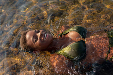Von oben fröhliche sexy afroamerikanische Frau im Badeanzug liegend mit geschlossenen Augen im Wasser des Meeres im Sommer - ADSF29550