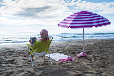 Back view of anonymous female freelancer sitting in lounger and working on tablet on sandy beach near sea in summer - ADSF29551