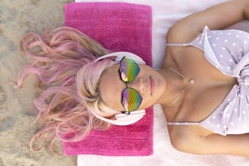 Draufsicht auf eine Frau mit rosa Haaren und Sonnenbrille, die auf einem Handtuch am Sandstrand liegt und sich im Sommerurlaub entspannt, während sie über Kopfhörer Musik hört - ADSF29553