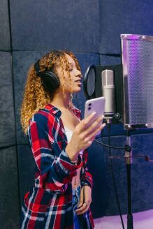 Fröhliche afroamerikanische Sängerin mit Kopfhörern und Smartphone, die in einem Aufnahmestudio ein Lied gegen den Schallschutz vorträgt - ADSF29559