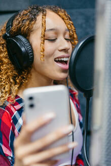 Fröhliche afroamerikanische Sängerin mit Kopfhörern und Smartphone, die in einem Aufnahmestudio ein Lied gegen den Schallschutz vorträgt - ADSF29560