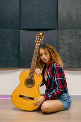 Young ethnic female guitarist with curly hair leaning on acoustic guitar while looking at camera - ADSF29565