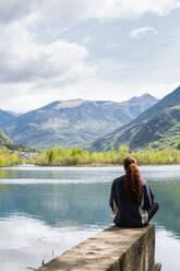 Back view of unrecognizable female traveler sitting on pier near pond and enjoying freedom in Pyrenees mountain range - ADSF29600