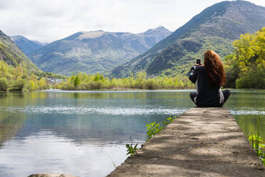 Rückenansicht einer nicht erkennbaren weiblichen Reisenden, die auf einem Steg sitzt und einen ruhigen Teich in den Pyrenäen fotografiert - ADSF29601