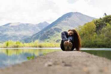 Female hiker sitting on stone quay near pond in Pyrenees mountain rage and looking at camera - ADSF29603