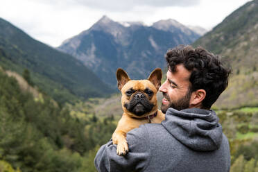Rückenansicht eines zufriedenen männlichen Wanderers, der auf einem Felsen sitzt und eine französische Bulldogge während eines Abenteuers in den Pyrenäen umarmt - ADSF29604