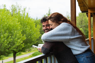 Side view of delighted couple of travelers hugging on terrace of wooden cottage and browsing mobile phone together during vacation in Pyrenees mountain range - ADSF29607