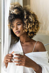 Friedliche Afroamerikanerin mit lockigem Haar und weißem Hemd, die mit einer Tasse Getränk am Morgen am Fenster steht - ADSF29610