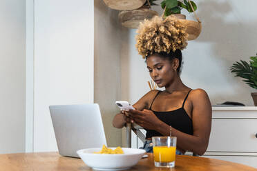 Attraktive afroamerikanische Freiberuflerin, die auf einem Stuhl sitzt und auf ihren Laptop schaut, während sie am Tisch beim Frühstück auf ihrem Smartphone surft - ADSF29613