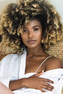 Delicate attractive African American female with curly hair sitting on sofa and touching skin while looking at camera - ADSF29617