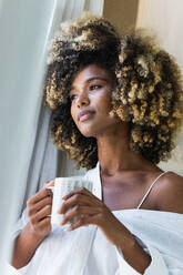 Friedliche Afroamerikanerin mit lockigem Haar und weißem Hemd, die mit einer Tasse Getränk am Morgen am Fenster steht - ADSF29618