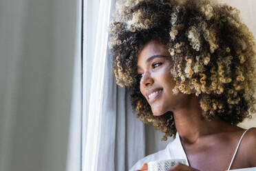 Friedliche, erfreute Afroamerikanerin mit lockigem Haar und weißem Hemd, die mit einer Tasse am Morgen am Fenster steht - ADSF29619