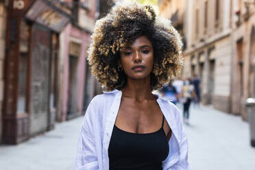 Confident African American female with blond Afro wearing top and white shirt standing between buildings and looking at camera - ADSF29621