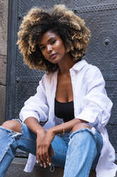 Confident African American female wearing stylish casual outfit sitting and looking at camera on concrete stairway against metal gates - ADSF29622