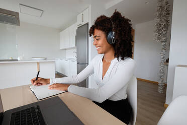 Lächelnde afroamerikanische Freiberuflerin mit Kopfhörern, die an einem Tisch mit Laptop sitzt und in einen Notizblock schreibt, während sie zu Hause an einem Projekt arbeitet - ADSF29638